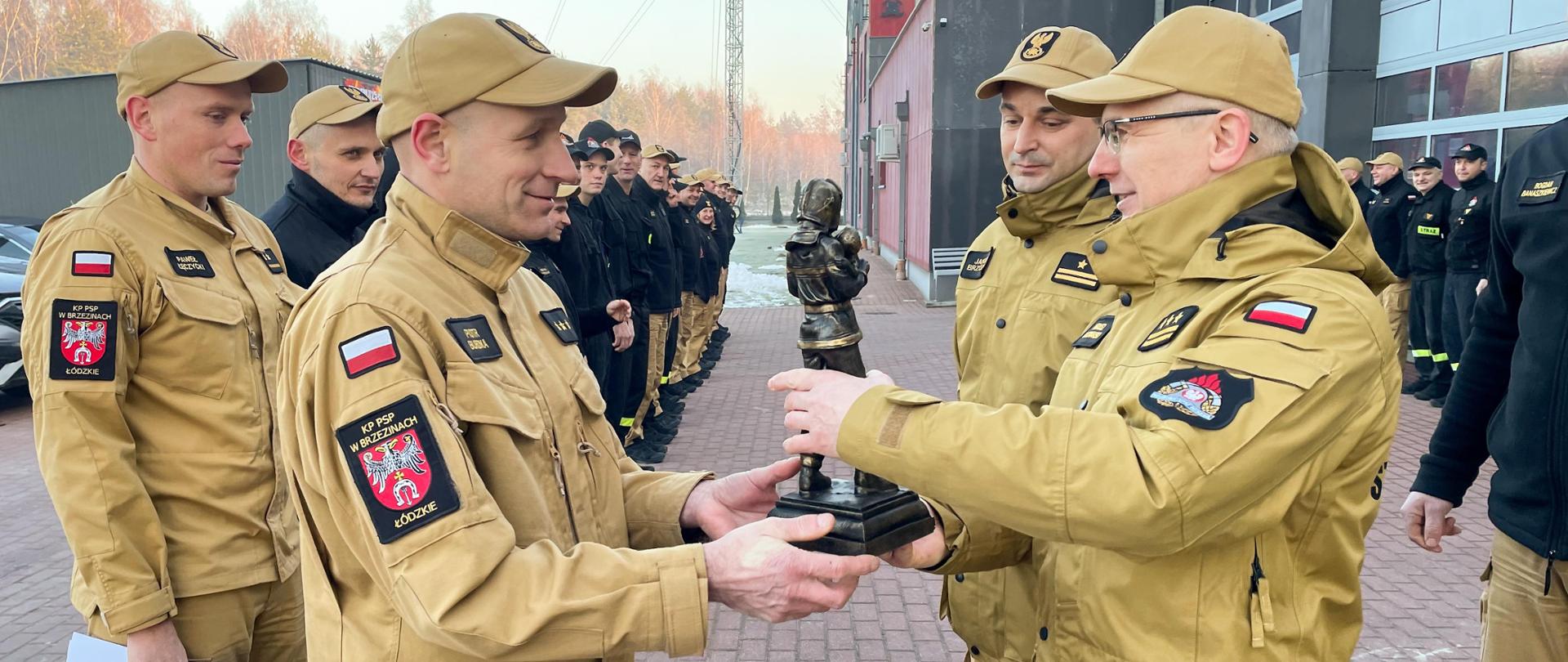 Przejście na zaopatrzenie emerytalne aspiranta sztabowego Piotra Burdki. Komendant Powiatowy Państwowej Straży Pożarnej w Brzezinach starszy brygadier Mariusz Kaczmarski wręcza pamiątkową statuetkę strażaka z okazji zakończenia służby przez aspiranta sztabowego Piotra Burdki. Podczas wręczania statuetki asystuje zastępca Komendanta Powiatowego Państwowej Straży Pożarnej w Brzezinach młodszy brygadier Jakub Brzózka. Sytuacja ma miejsce podczas uroczystej zbiórki na placu komendy, strażacy stoją w dwóch szeregach na przeciw siebie, 