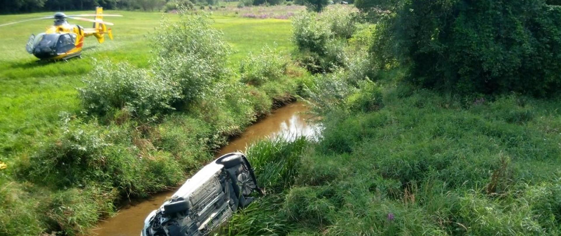 Zdjęcie przedstawia rzekę, w której na boku leży samochód osobowy, za nim, na pobliskiej łące wylądował śmigłowiec Lotniczego Pogotowia Ratunkowego..