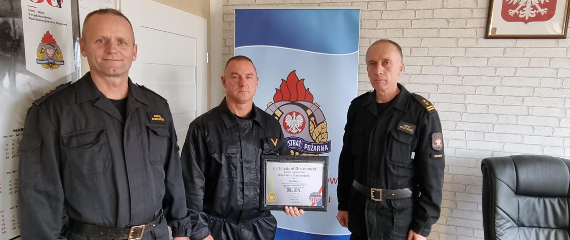 Zdjęcie przedstawia trzech strażaków w biurze komendanta, który pokazuje zdobyte trofeum.