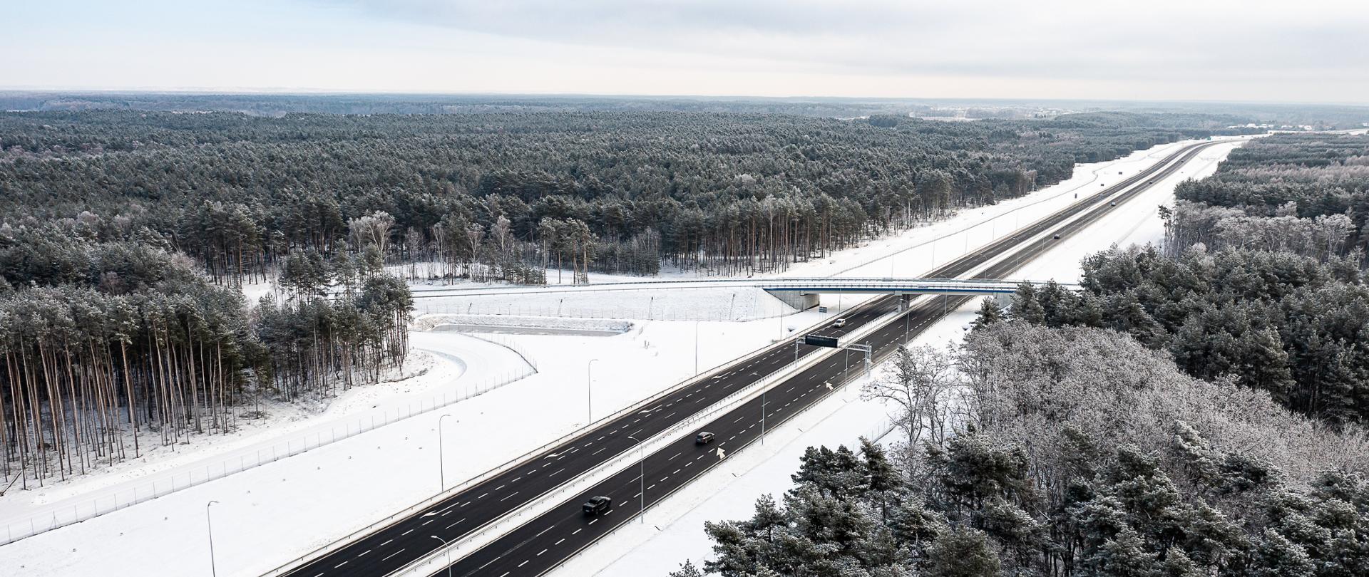 Sceneria zimowa, leży śnieg, widać drogę, która biegnie pomiędzy lasami. Droga odśnieżona. Widać wiadukt nad drogą.