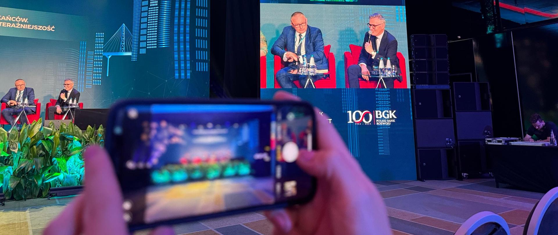 Na pierwszym planie dłonie trzymające smartfon, na ekranie scena z prelegentami, w tle ekran z bliskim ujęciem panelistów