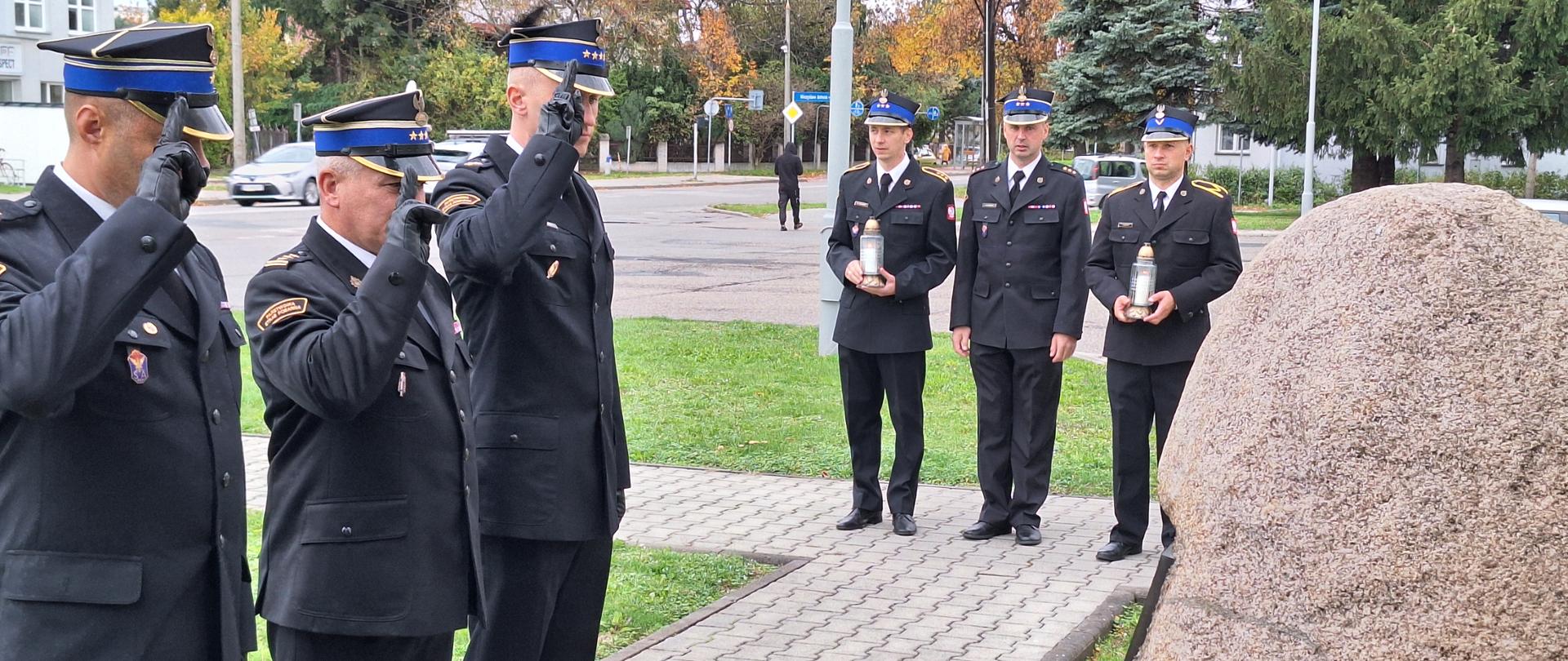 Strażacy ubrani w mundury galowe ustawieni w szeregu po 3 osoby salutują naprzeciw obeliska upamiętniającego tragiczną śmierć kolegów, obok druga trójka strażaków stoi ze zniczami