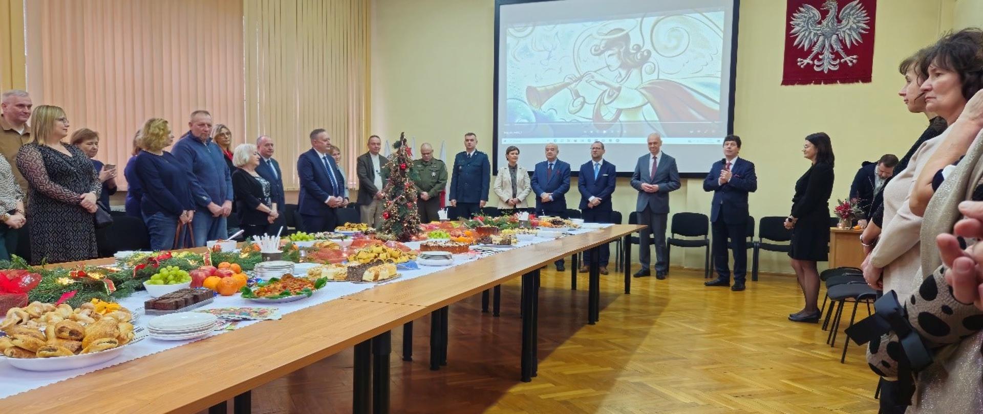 Zdjęcie przedstawia formalne zgromadzenie ludzi stojących w sali konferencyjnej. Długi stół na pierwszym planie jest zastawiony świątecznymi dekoracjami, jedzeniem i napojami, co sugeruje święto lub wydarzenie okolicznościowe. Na ścianie w tle znajduje się duży ekran wyświetlający artystyczną ilustrację postaci grającej na trąbce, a w pobliżu umieszczono czerwony emblemat z białym orłem. Uczestnicy są ubrani w formalne lub oficjalne stroje, a niektórzy wydają się reprezentować służby mundurowe.
