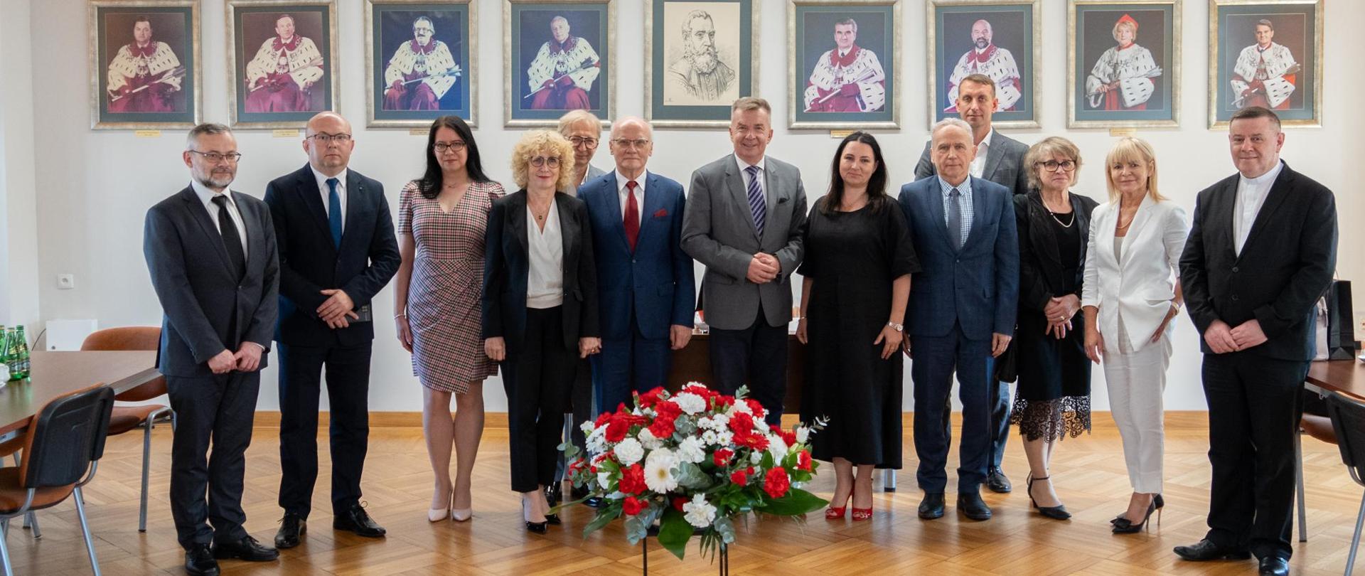 Zdjęcie zbiorowe, pod ścianą na której wisi rząd portretów stoi grupa trzynastu osób, pośrodku minister Wieczorek, przed grupą na podłodze bukiet biało-czerwonych kwiatów.