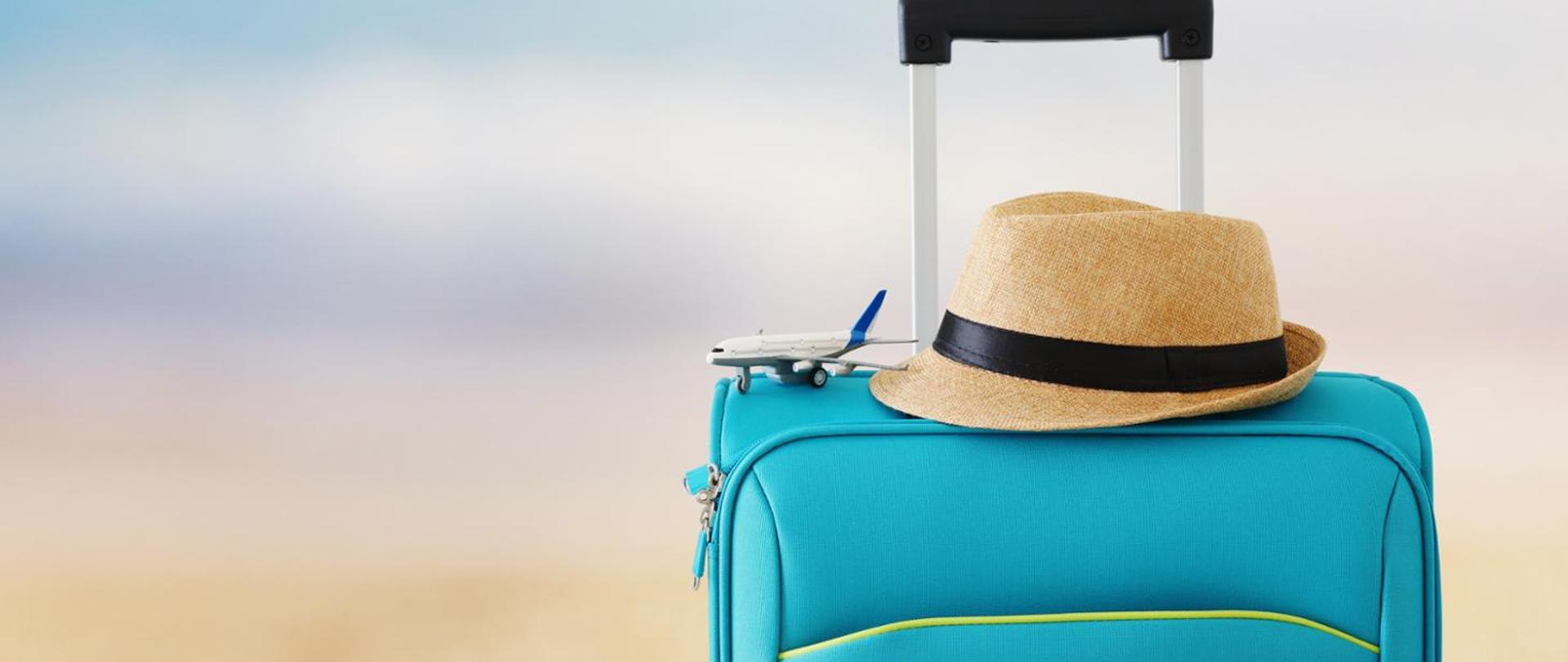 Po prawej stronie widać niebieską walizkę z wysuniętą rączką. Na walizce lezy miniaturka samolotu oraz kapelusz przeciwsłoneczny.