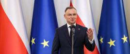 Prezydent Duda stoi i mówi do mikrofonu na stojaku, za nim rząd flag Polski i UE.