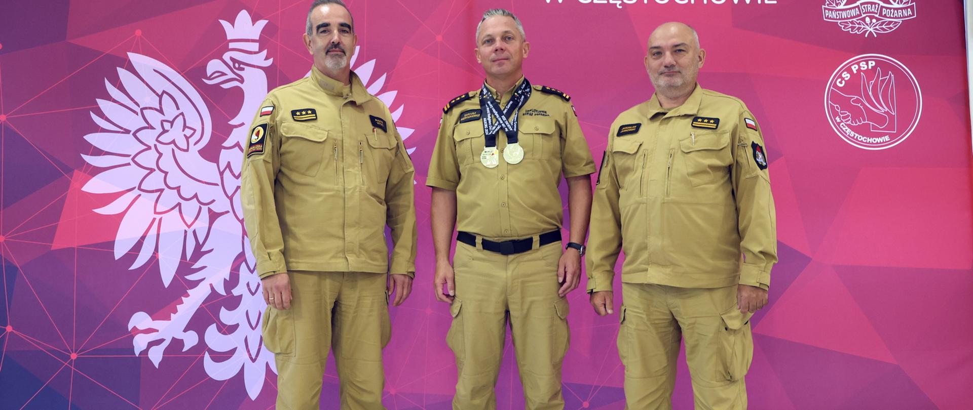 Pamiątkowe zdjęcie funkcjonariusza CS PSP, będącego uczestnikiem XV Światowych Igrzysk Strażaków World Firefighters Games 2024, z dwoma złotymi medalami na szyi, w towarzystwie Komendanta Centralnej Szkoły PSP oraz Zastępcy Komendanta CS PSP na tle baneru mającego u góry napis Centralna Szkoła Państwowej Straży Pożarnej w Częstochowie, po prawej logo PSP, poniżej logo CS PSP pod napisem duży orzeł w koronie