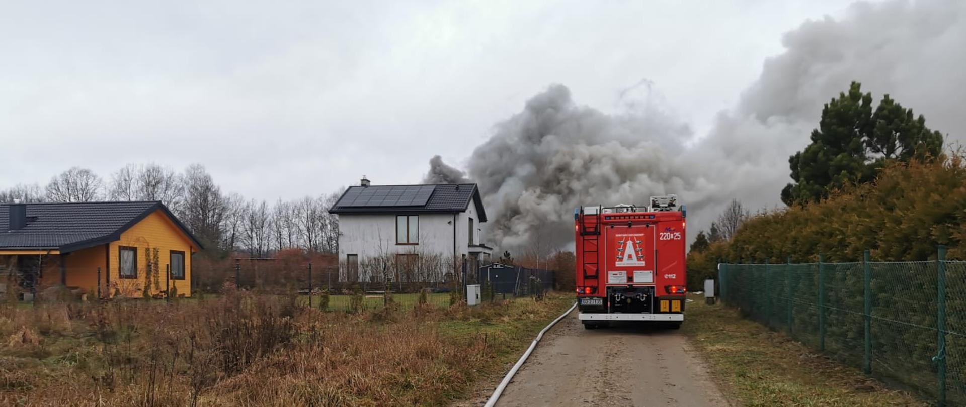 Samochód strażacki na tle kłębów dymu unoszących się nad objętym pożarem garażem.