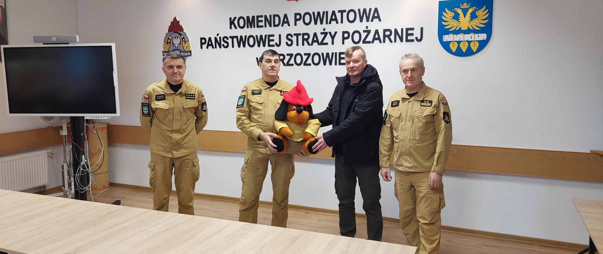 Kolorowa fotografia wykonana w pomieszczeniu świetlicy Komendy Powiatowej PSP w Brzozowie. Przedstawia trzech strażaków w umundurowaniu służbowym oraz osobę cywilną, której przekazana jest maskotka strażacka tzw. Żarek w ramach WOŚP.