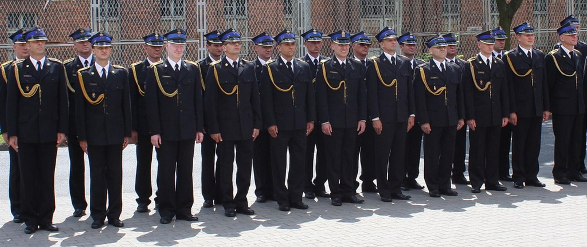 Zdjęcie przedstawia stojących w szeregu strażaków z KP PSP Pleszew ubranych w mundury wyjściowe