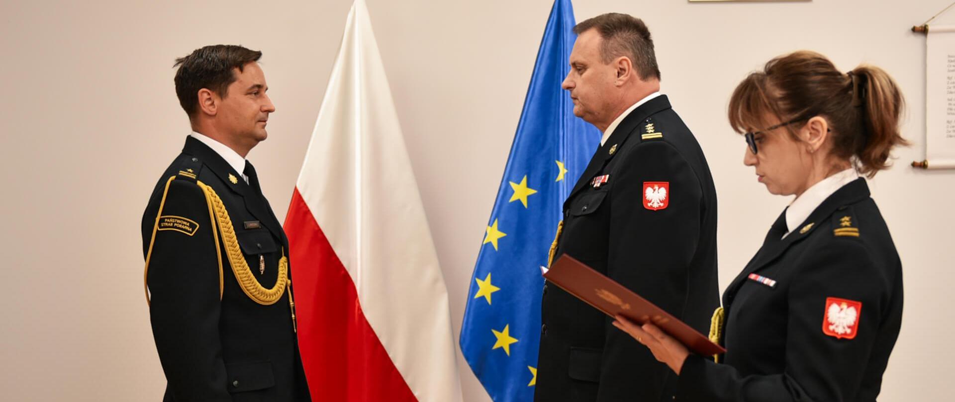 po lewej stoi jednej strażak na przeciwko niego dwóje strażaków, funkcjonariuszka stojąca po prawej trzyma w rękach teczkę i odczytuje pismo, za nimi flagi polski i unii europejskiej