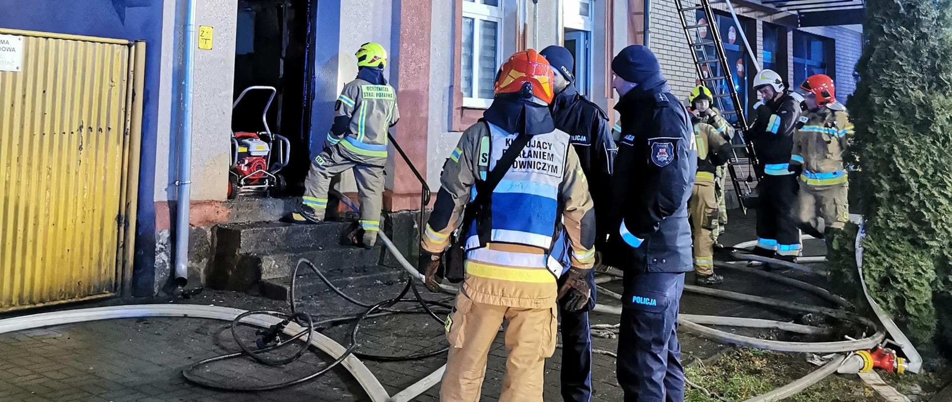 Zdjęcie przedstawia strażaków podczas gaszenia pożaru kamienicy.
W tle budynek.
