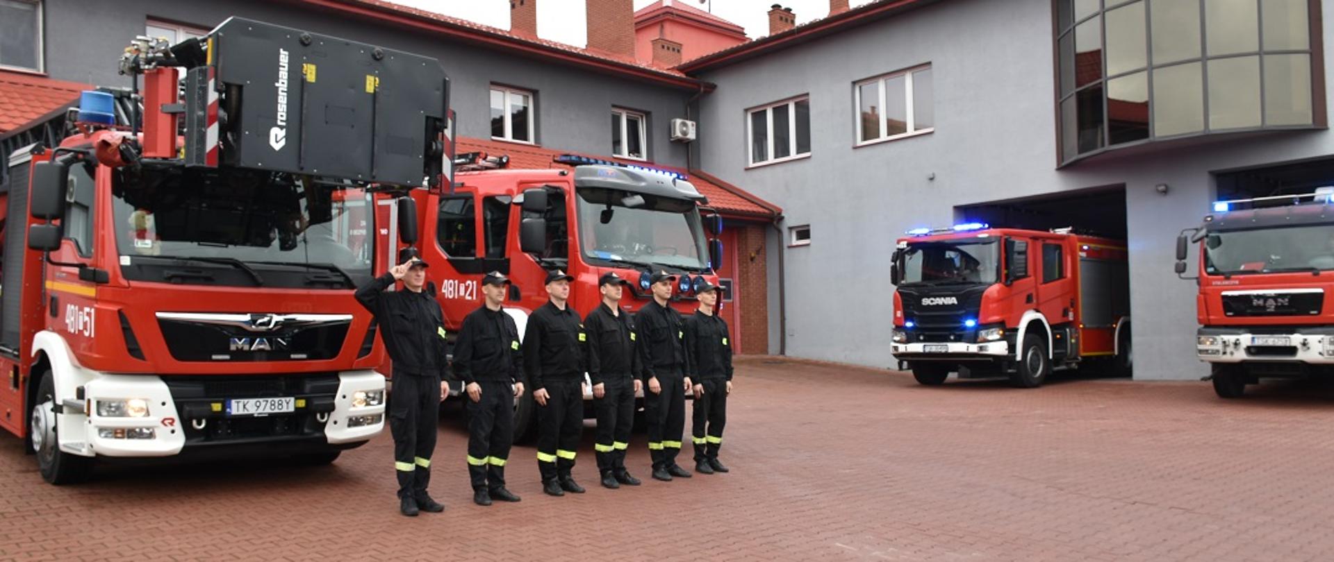 Na fotografii na pierwszym planie widzimy stojących w szeregu strażaków ubranych w ubrania specjalne koloru czarnego. Strażacy stoją w postawie zasadniczej. Jeden ze strażaków stojący na początku szeregu oddaje salut. W tle widać dwa stojące pojazdy pożarnicze koloru czerwonego oraz budynki komendy powiatowej PSP w Skarżysku-Kamiennej. W pojazdach zostały załączone sygnały świetlne.