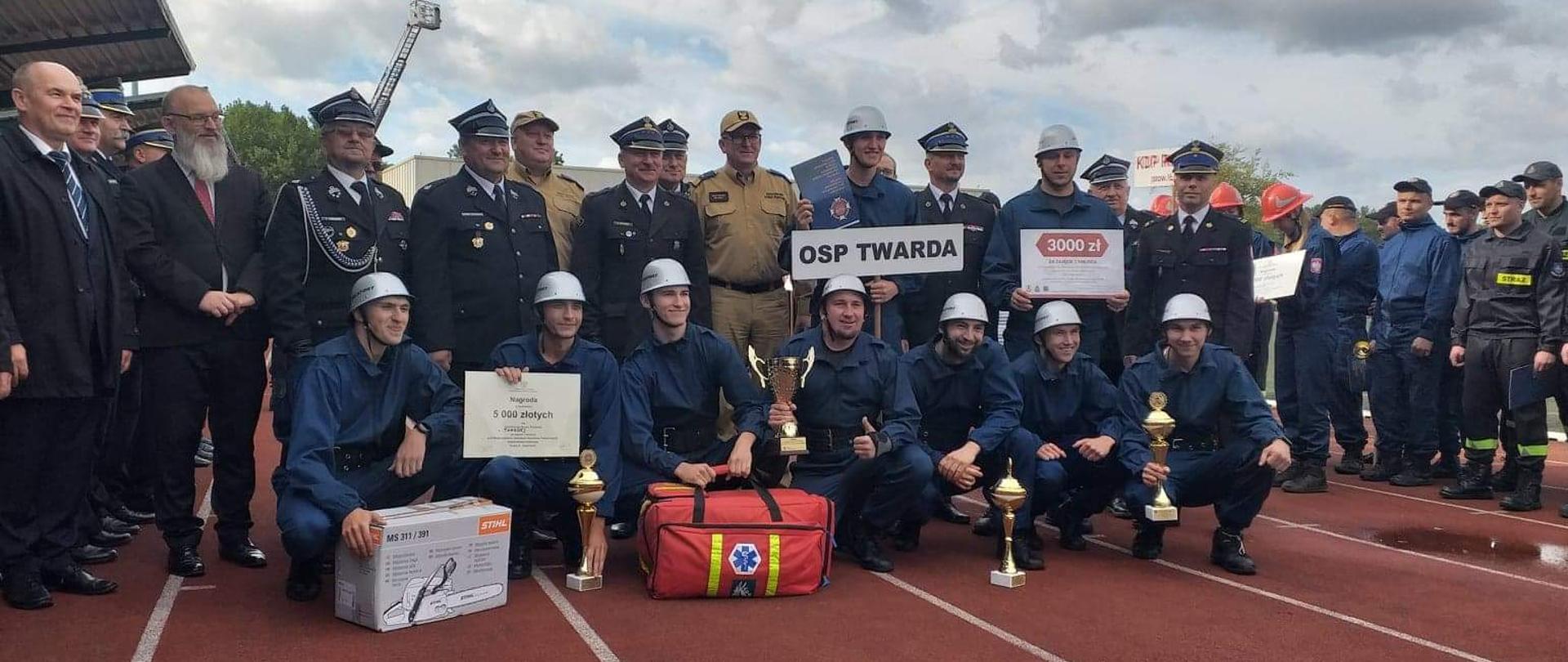 Na pierwszym planie widać męską drużynę pożarniczą z OSP Twarda. Za nimi stoją strażacy. Wszyscy stoją na bieżni tartanowej. Mężczyźni z OSP Twarda trzymają nagrody.