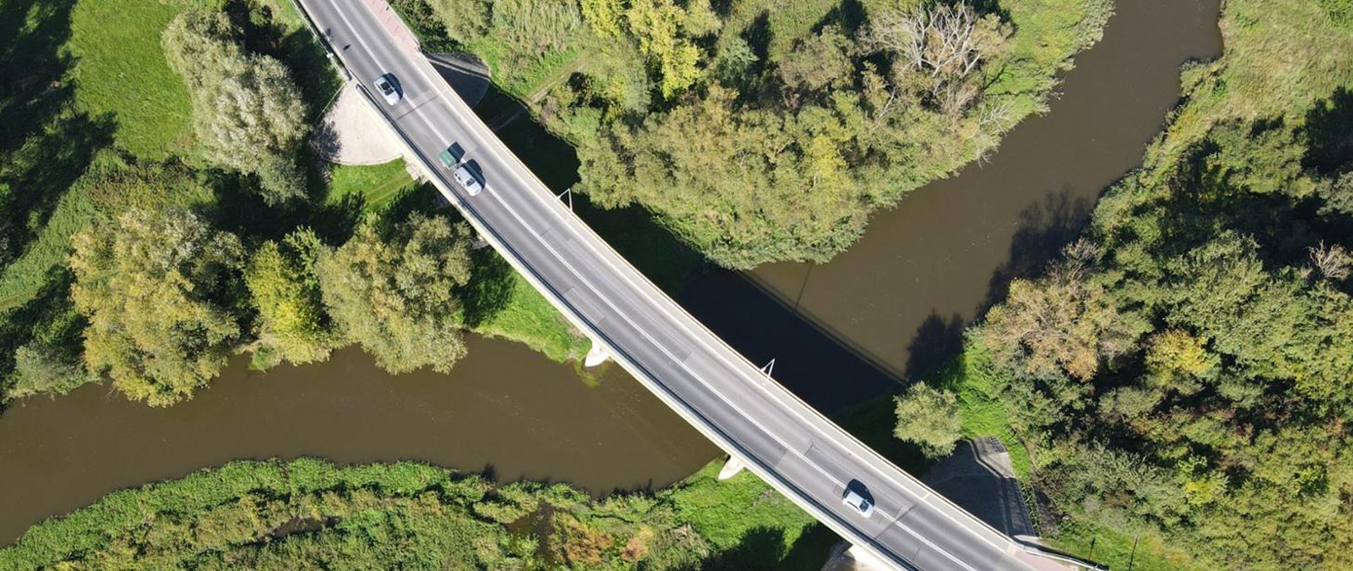 Widok z lotu ptaka na most biegnący nad rzeką. Na moście widoczne pojazdy, na brzegach rzeki bujna roślinność.