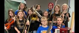 Grupa uczniów w czarnych i niebieskich koszulkach z logo szkoły trzymających instrumentu muzyczne.