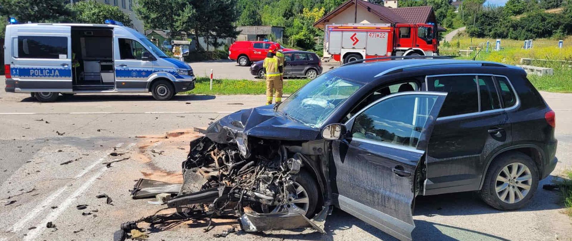 Zdjęcie przedstawia rozbity samochód na jezdni w tle samochody straży pożarnej oraz radiowóz Policji.