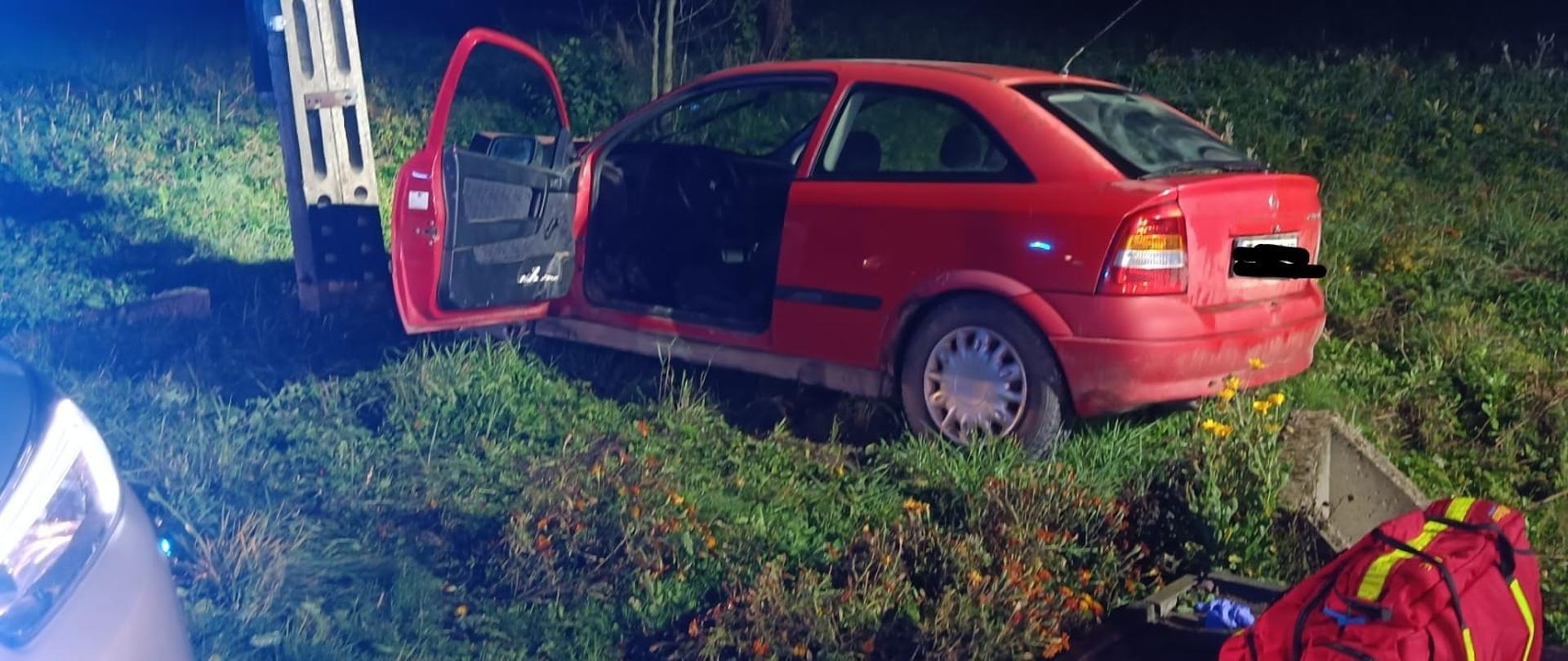 Samochód osobowy koloru czerwonego uderzył w przydrożnego słupa, drzwi od kierowcy otwarte, na dole po prawej stronie stoi torba medyczna. Na zdjęciu jest ciemno