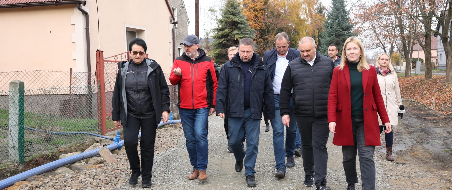 Ministrowie razem z Wojewodą oraz pozostałymi uczestnikami odwiedzili Bodzanów. Idą drogą obok domów. 