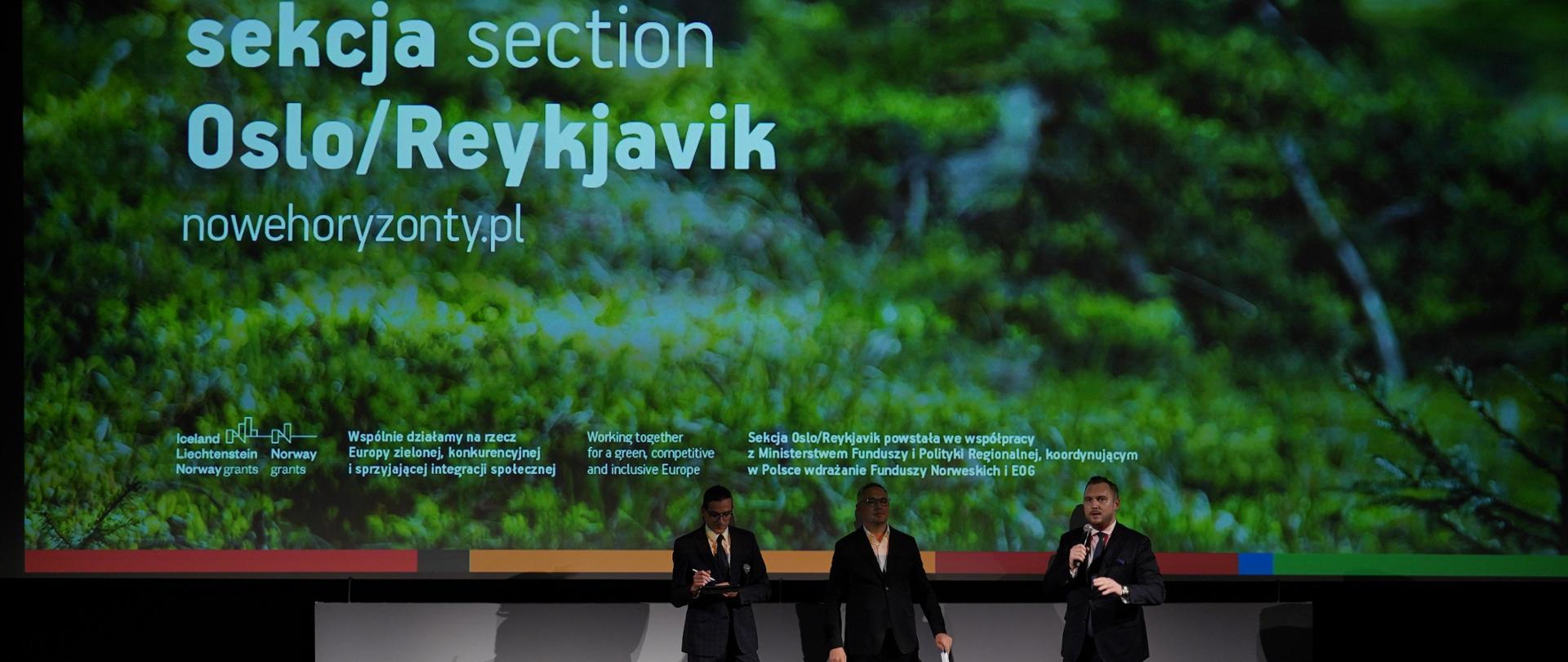 Trzy osoby stoją obok siebie na scenie. Pierwszy od prawej z mikrofonem w prawej dłoni stoi wiceminister Konrad Wojnarowski. 