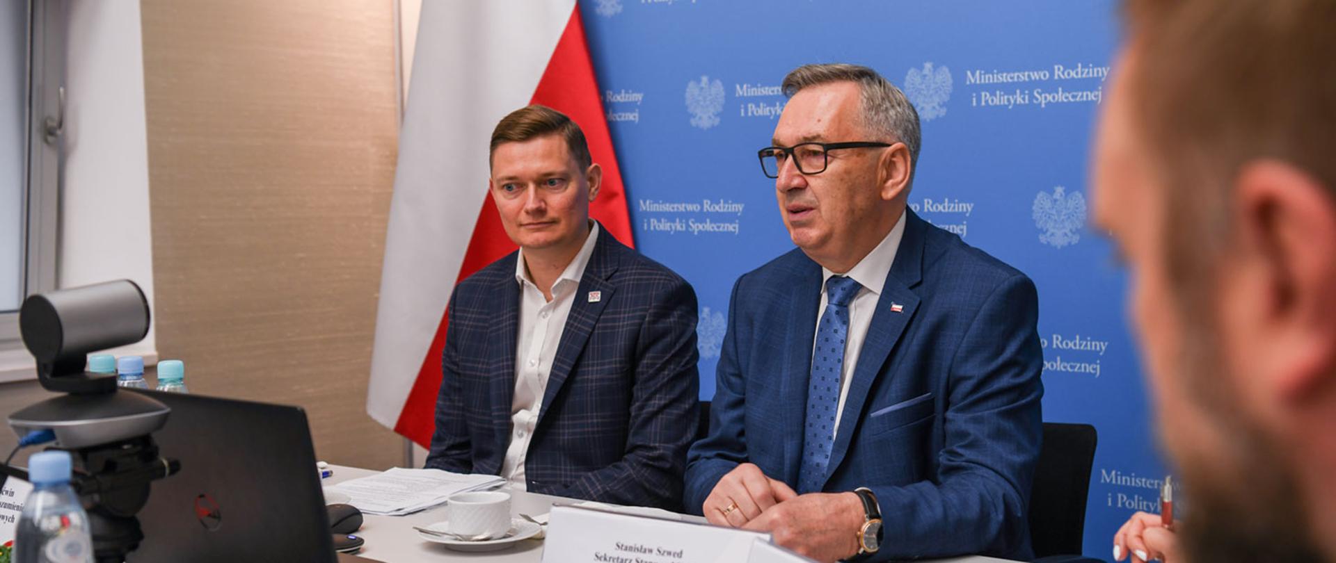 Zdjęcie przy stole. Widoczni dwaj mężczyźni. Jeden z nich to minister Szwed. W tle niebieski baner z logiem Ministerstwa Rodziny i Polityki Społecznej.