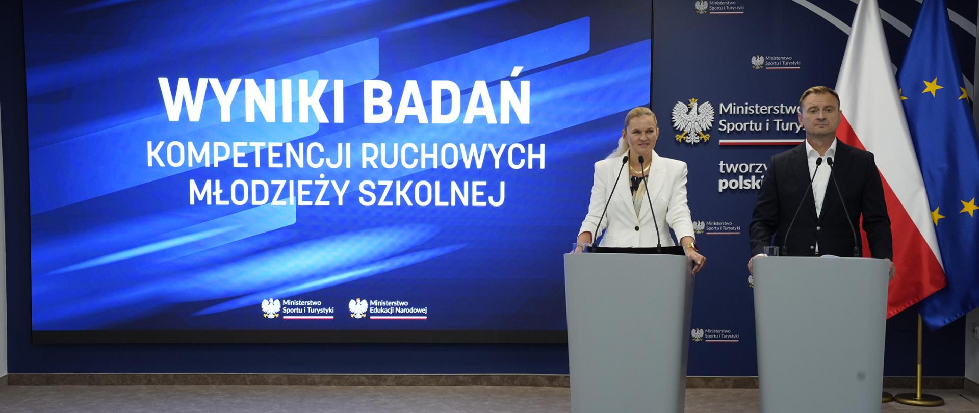 Na tle dużego ekranu na którym wyświetlany jest tekst: WYNIKI BADAŃ KOMPETENCJI RUCHOWYCH MŁODZIEŻY SZKOLNEJ" po prawej stroni stoi minister Barbara Nowacka i minister Sławomir Nitras. Na ekranie wyświetlane są również loga Ministerstwa Sportu i Turystyki oraz Ministerstwa Edukacji Narodowej. W tle znajduje się również flagi Polski i UE