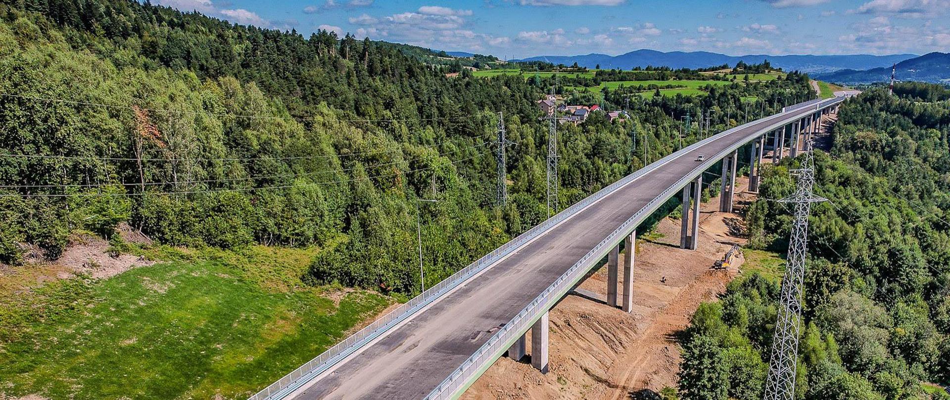 Na zdjęciu widzimy widok z lotu ptaka na długą estakadę. Obiekt jest podtrzymywany przez wiele filarów i znajduje się na obszarze otoczonym zielenią, w tym lasami i otwartymi polami. Na wiadukcie znajduje się jeden pojazd. Niebo jest częściowo zachmurzone, a w oddali widać góry na horyzoncie.