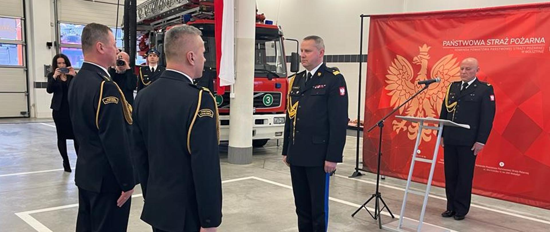 w garażu dwóch strażaków stoi na baczność przed jednym strażakiem, generałem, w tle mównica i czerwony baner z orłem oraz samochód strażacki