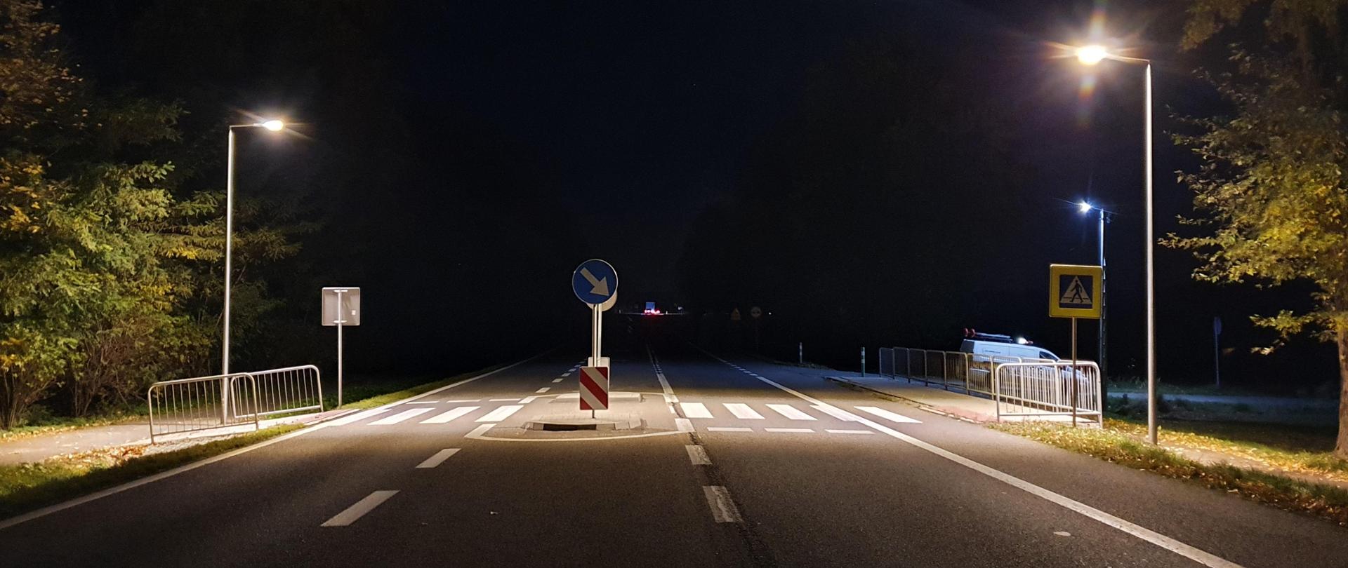 Oświetlone przejście dla pieszych. Zdjęcie robione w późnych godzinach nocnych. W oddali widoczne pojazdy.