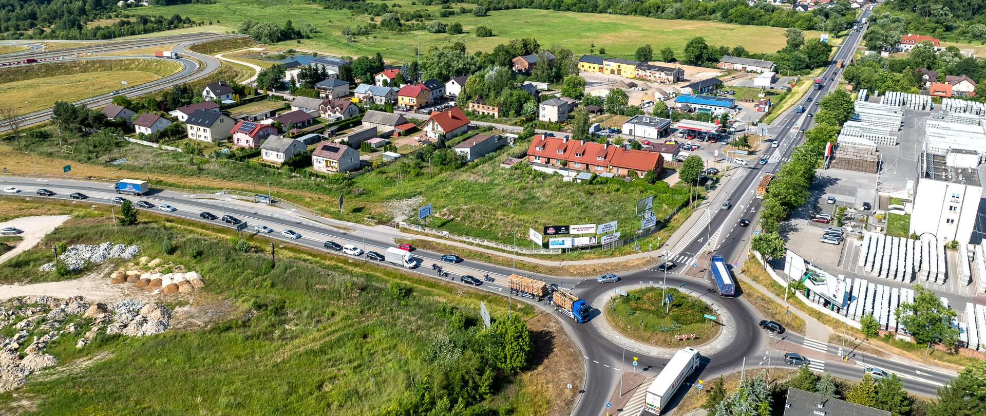 Na zdjęciu widać widok z lotu ptaka na skrzyżowanie dróg z rondem. Widać wiele pasów ruchu oraz poruszające się pojazdy. Wokół skrzyżowania znajdują się budynki, a także kilka domów mieszkalnych. Obszar otaczają drzewa i zieleń, co nadaje mu przyjemny wygląd. Widać również prace budowlane w pobliżu ronda. Niebo jest czyste z kilkoma chmurami.