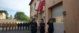 Na pierwszym planie trzech strażaków w umundurowaniu galowym: dwóch z nich salutuje, trzeci z nich (w środku) wciąga flagę na maszt. Z tyłu za nimi w szeregu stoją strażacy w umundurowaniu koszarowym. Po prawej stronie budynek z dużymi bramami garażowymi i oknami w górnej części. Po prawej w tle mniejsze budynki, za nimi drzewa.