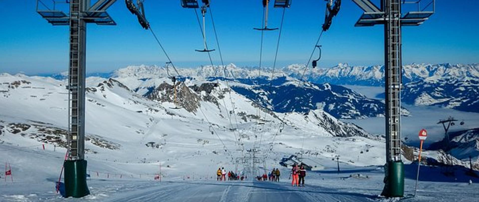 Na zdjęciu widnieje ośnieżony stok narciarski z wyciągiem, w tle panorama gór.
