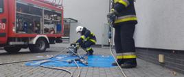 Strażacy ochotnicy z OSP Tuchorza w trakcie zdawania egzaminu praktycznego. Na pierwszym planie strażak obsługuje narzędzie hydrauliczne. W tle, drugi strażak zajmuje się obsługą pompy hydraulicznej.