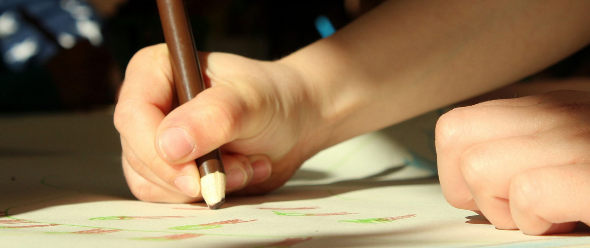 Obraz przedstawia rękę trzymającą brązową kredkę w trakcie rysowania pracy na papierze