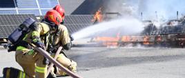 Dwaj strażacy widoczni z tyłu z wężem strażackim leją wodę na palące się panele fotowoltaiczne. Widoczne płomienie. Z lewej ztrony zdjęcia ustawiona instalacja fotowoltaiczna nieobjęta pożarem.