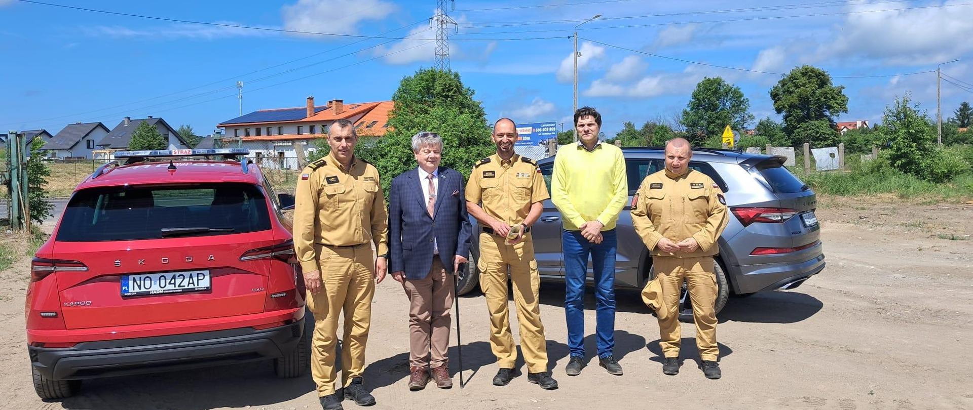 Na zdjęciu piećiu osób, trzech funkcjonariuszy Państwowej straży Pożarnej. W środku stoi Komendant Wojewódzki oraz starosta braniewski. Z lewej strony stoi czerwony samochód. W tle niebieskie niebo, słonecznie.