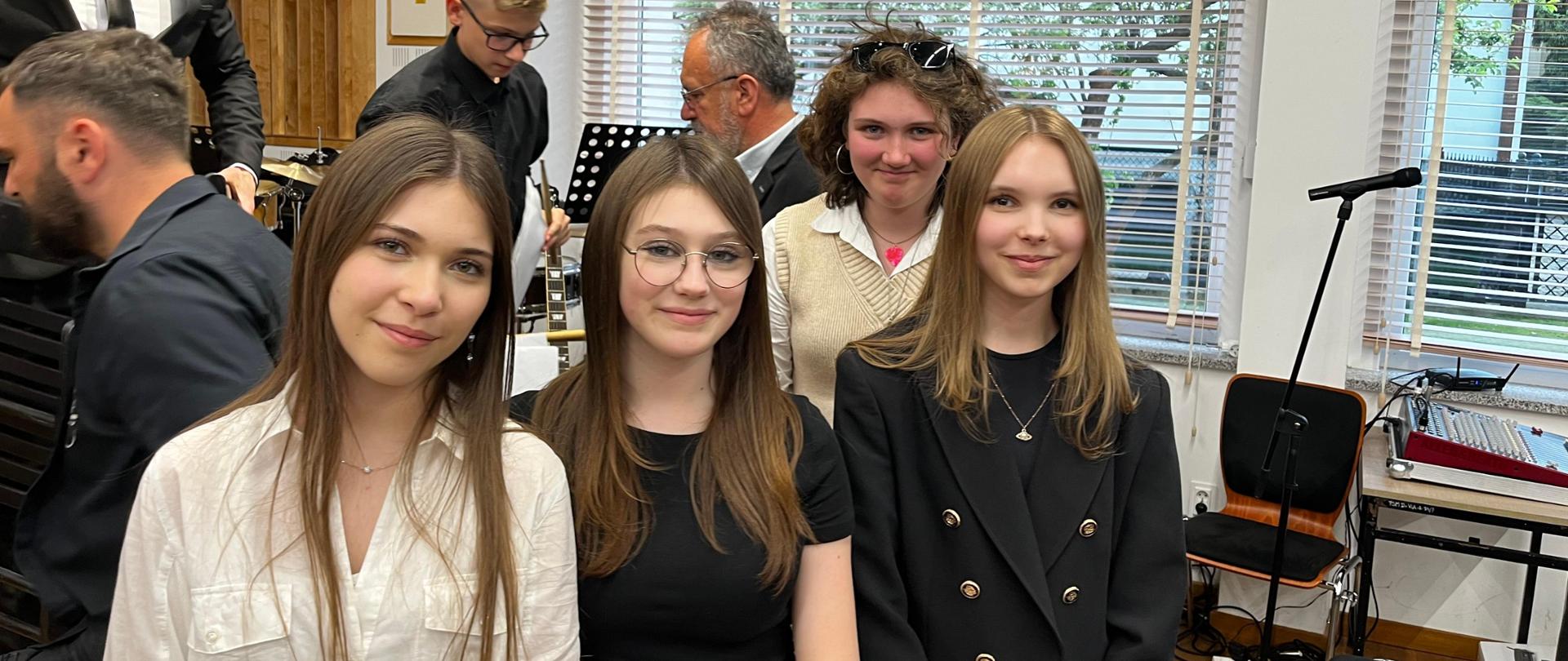 Grupa uśmiechniętych uczennic w strojach galowych stoi w jednej z klas PSM w Gorlicach. W tle pedagog i inni uczniowie. Ogólna kolorystyka czarno-brązowa.