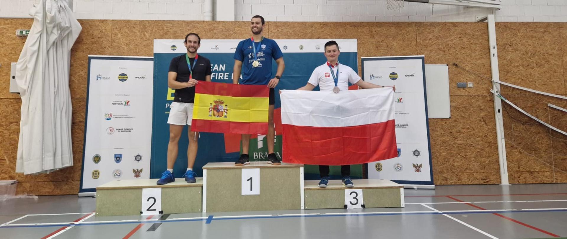 Mł. kpt. Michał Stebnicki na podium. 3 miejsce. Trzyma Flagę Polski. Na 1 i 2 miejscu mężczyźni trzymają flagę Hiszpanii