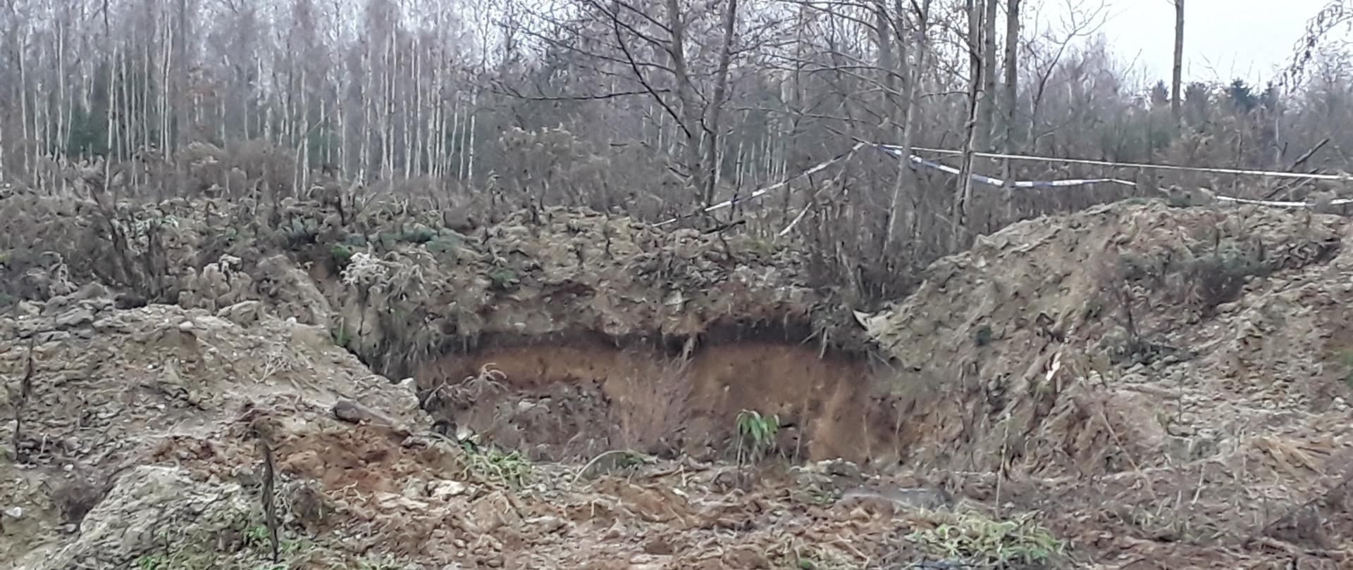 Wykop w ziemi o powierzchni kilku metrów kwadratowych na otwartym terenie, wcześniej przygotowany do zakopania odpadów, które zostały usunięte. W tle las.