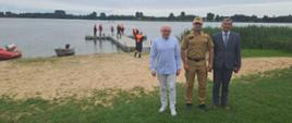 Zdjęcie przedstawia plażę gminną w Głuszynie. Na pierwszym planie Wójt Gminy Bytoń, Sekretarz Gminy Bytoń oraz Komendant Powiatowy PSP w Radziejowie. Na drugim planie strażacy PSP oraz OSP podczas ćwiczeń na wodzie jeziora Głuszyńskiego. 