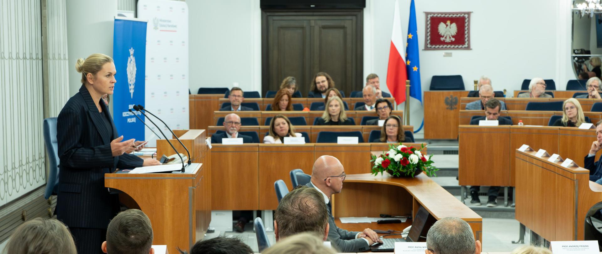 Minister Edukacji Barbara Nowacka przemawia z mównicy w Senacie. Na sali siedzą przysłuchujący się wypowiedzi uczestnicy konferencji edukacyjnej. W tle ustawiona jest flaga Polski oraz Unii Europejskiej, a na ścianie wisi godło polskie. 