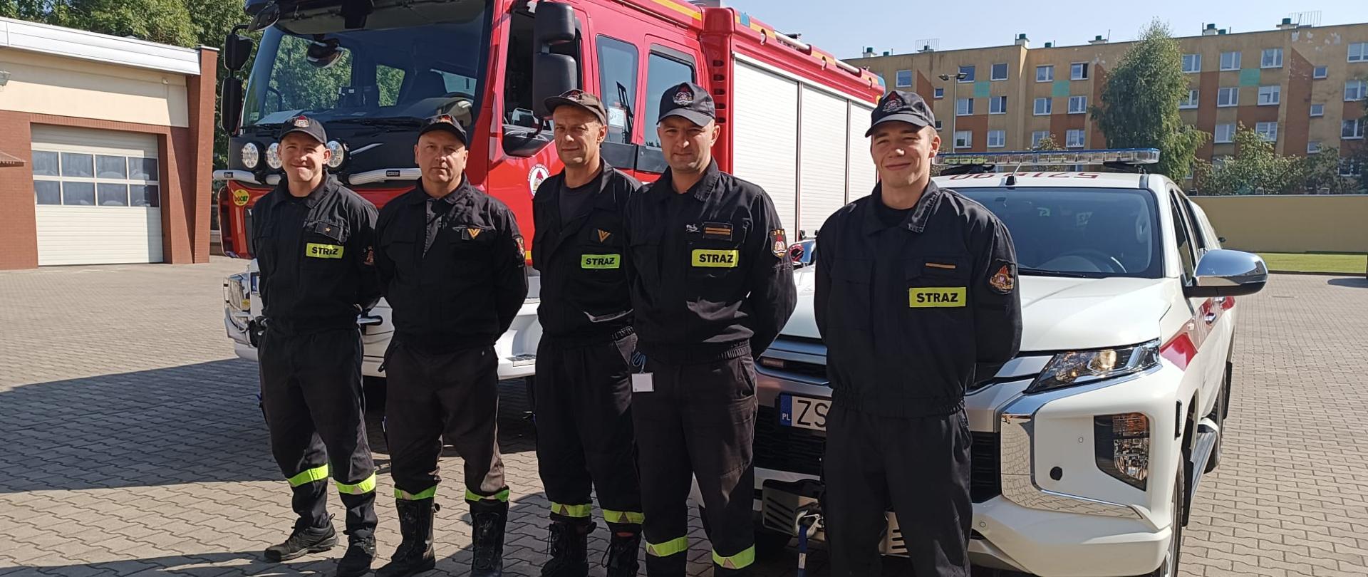 Na zdjęciu znajduje się pięciu wałeckich strażaków wyznaczonych do wyjazdu do działań związanych z usuwaniem skutków powodzi na południu Polski. Strażacy ustawieni w szeregu. W tle ustawione dwa pojazdy pożarnicze.