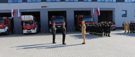 Dzień strażaka przed JRG 1 Kielce. Strażacy na zbiórce przed jednostką. Składany jest meldunek przez strażaka w piaskowym ubraniu strażakowi w mundurze wyjściowym.