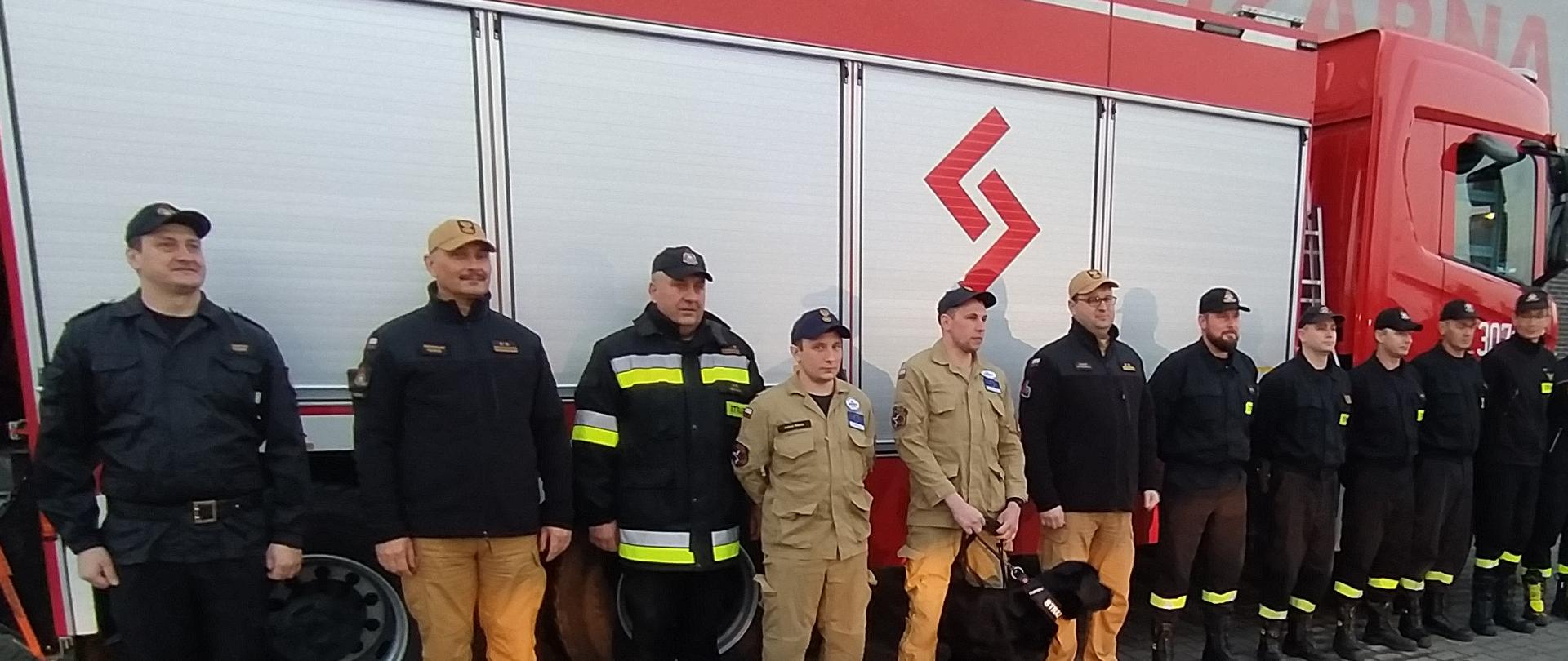 grupa strażaków stoi na bacznoś przed samochodem strażackim