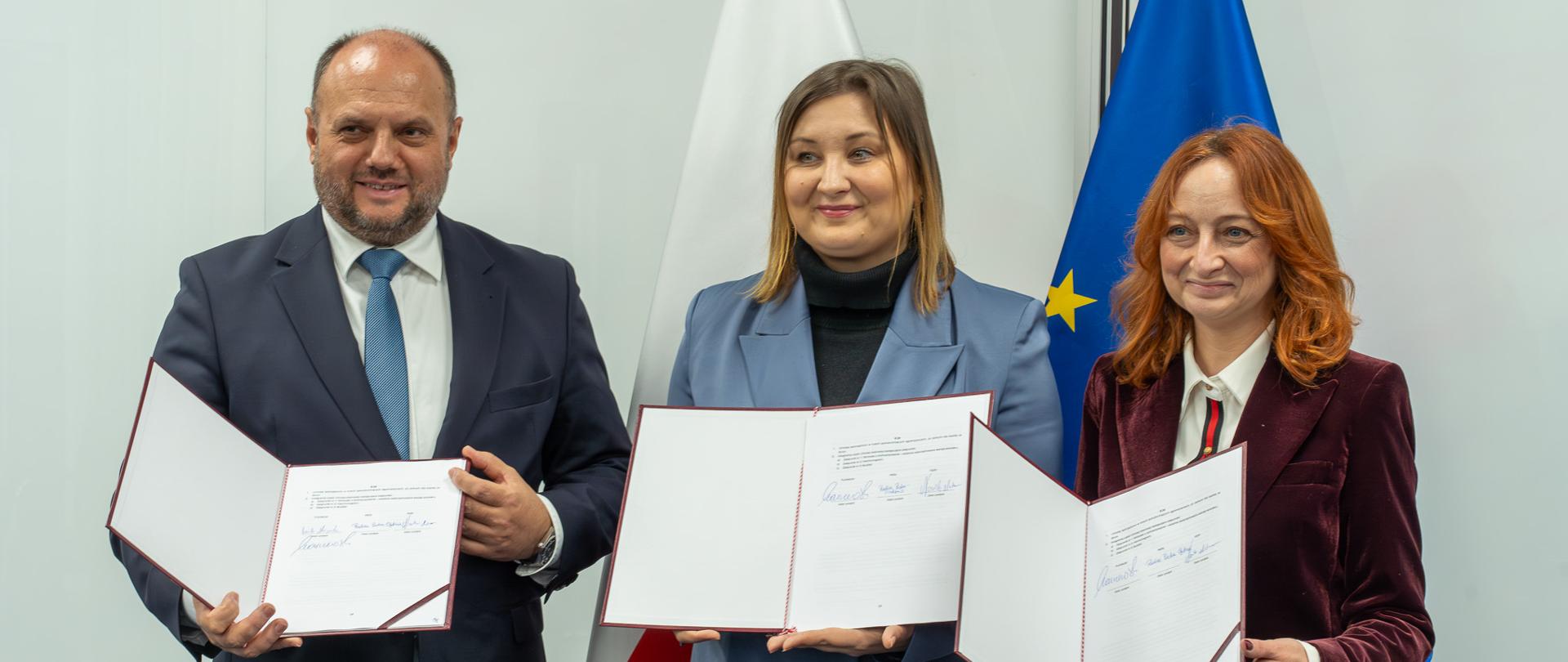 Widok z boku. Na tle flag Polski i UE stoi trzy osoby w środku wiceminister Paulina Piechna-Więckiewicz i każda z nich trzyma otwarty dokument, który przed chwilą podpisała.