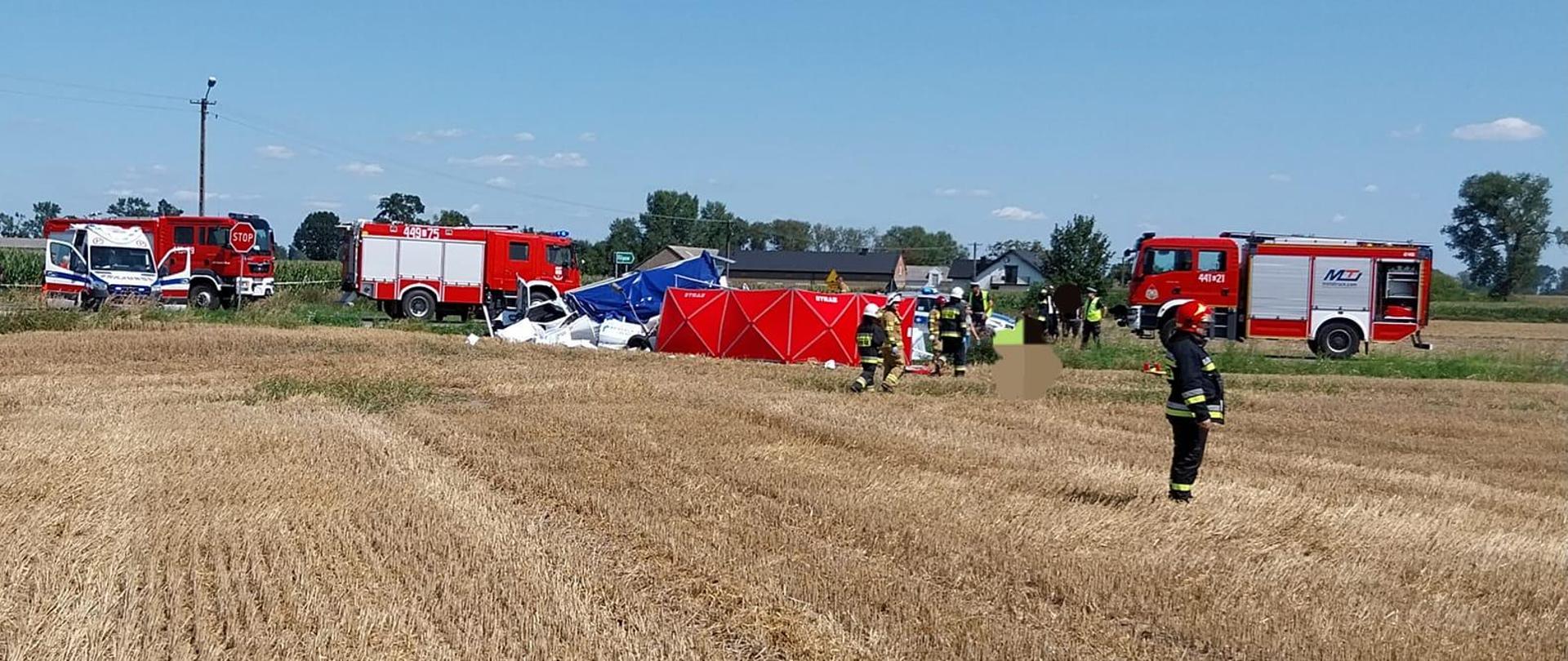 Na zdjęciu z oddali widać rozbity samochód dostawczy z białą kabiną i niebieską plandeką, trzy czerwone samochody strażackie, karetkę pogotowia, strażaków, policjantów, budynki oraz drzewa. Na polu po skoszonym zbożu stoi strażak w ciemnym stroju i czerwonym hełmie. Jest widno.