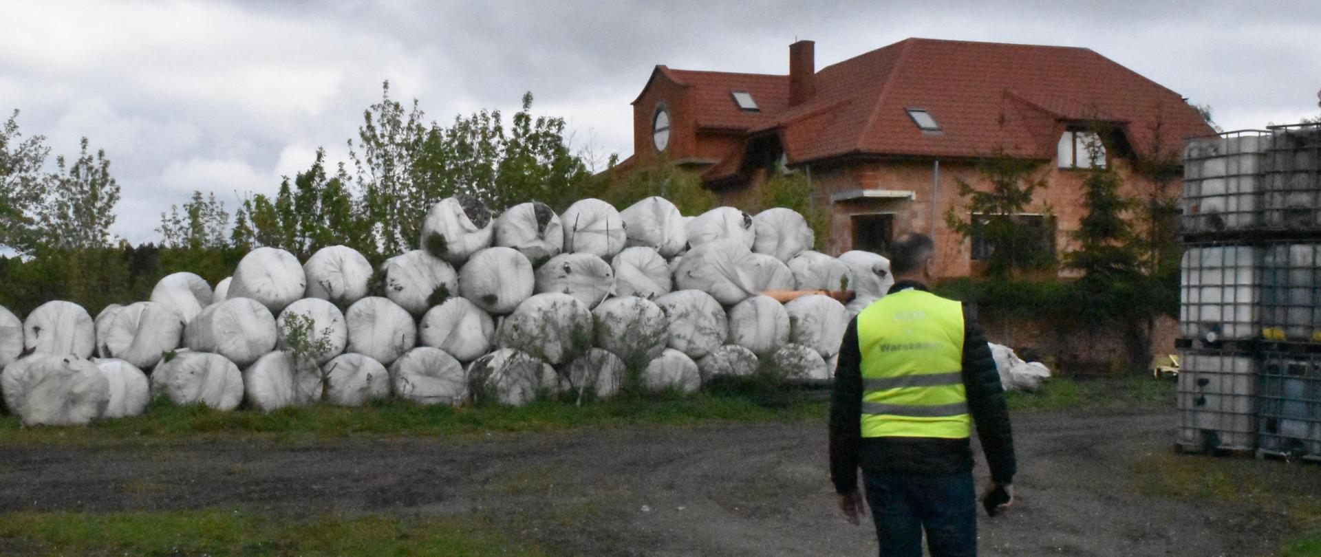 Inspektor Wojewódzkiego Inspektoratu Ochrony Środowiska prowadzi wizję terenową miejsca nielegalnego gromadzenia odpadów. Po prawej stronie stoją ustawione w trzech warstwach pojemniki typu mauzer. Na wprost leżą bele o średnicy 1,2 m z odpadami komunalnymi. W tle widać jednopiętrowy budynek.