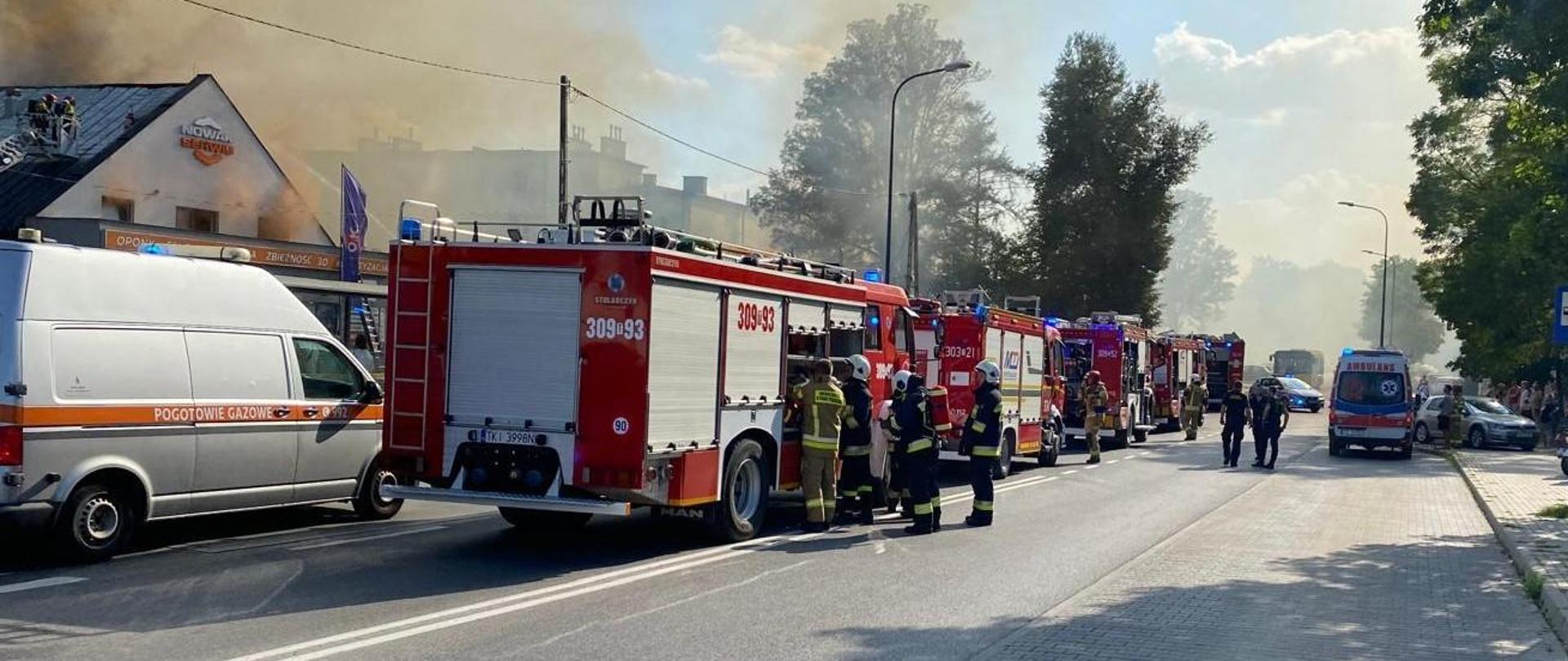 Ulica Wojciecha Szczepaniaka i zaparkowane na niej samochody służb ratowniczych. Kolejno od lewej stoją: Pogotowie Gazowe, 5 samochodów gaśniczych, po prawej ambulans ZRM a w głębi widać policyjny radiowóz. Na jezdni, między pojazdami przemieszczają się funkcjonariusze.