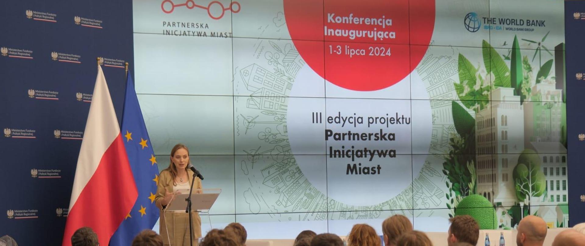 Minister Katarzyna Pełczyńska-Nałęcz stoi w oddali na scenie przy pulpicie i mówi do mikrofonu do zgromadzonych na sali osób. 