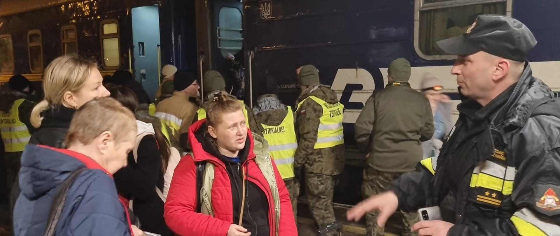 Zdjęcie przedstawia strażaka w ubraniu dowódczo-sztabowym, który znajduje się na peronie kolejowym i w ręku trzyma telefon. Wokół niego zgromadzonym kilkanaście uchodźców wojennych, którzy przybyli pociągiem z Ukrainy.